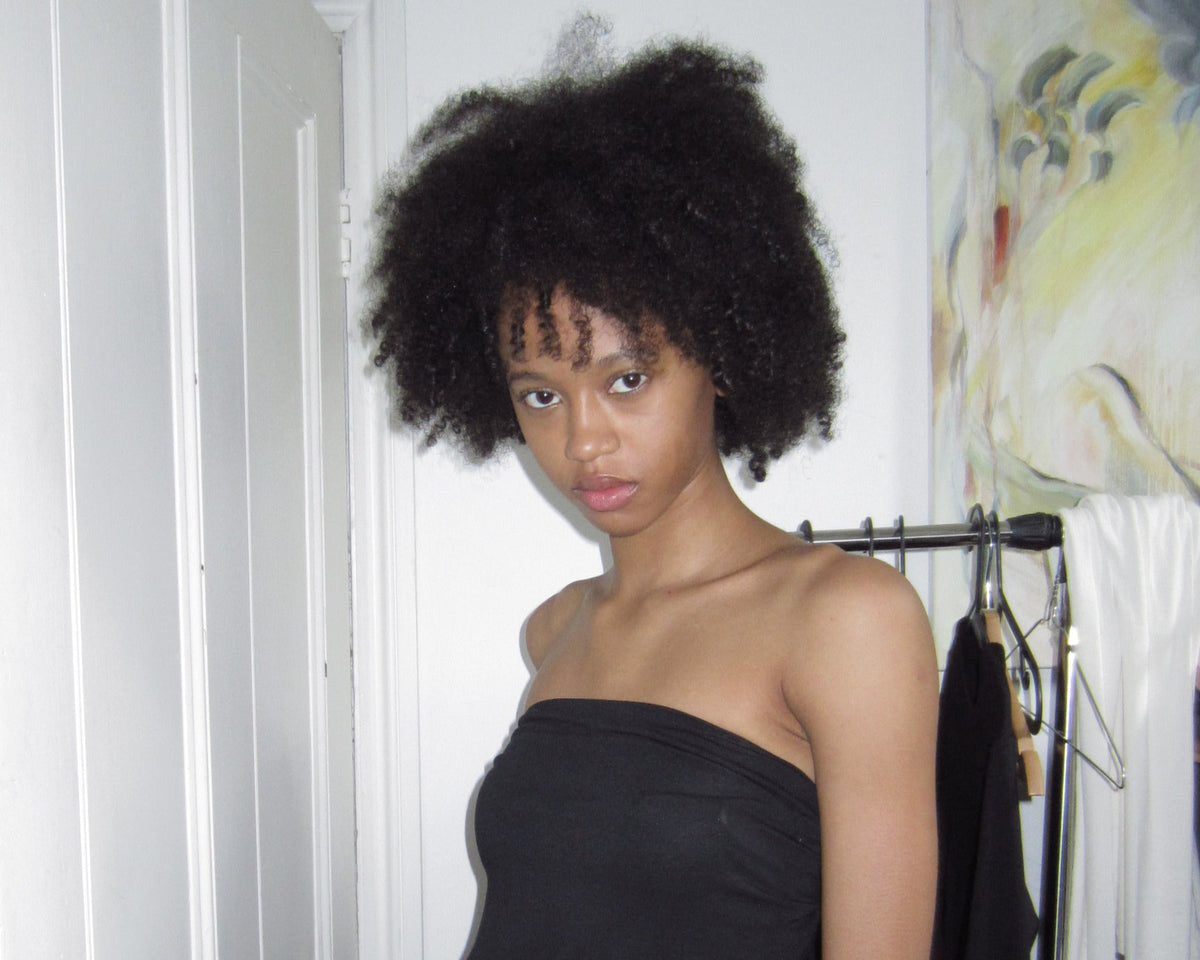 A woman with a large black afro hairstyle wearing a black strapless dress with a side cutout, standing in a room with a white door and a clothing rack in the background.